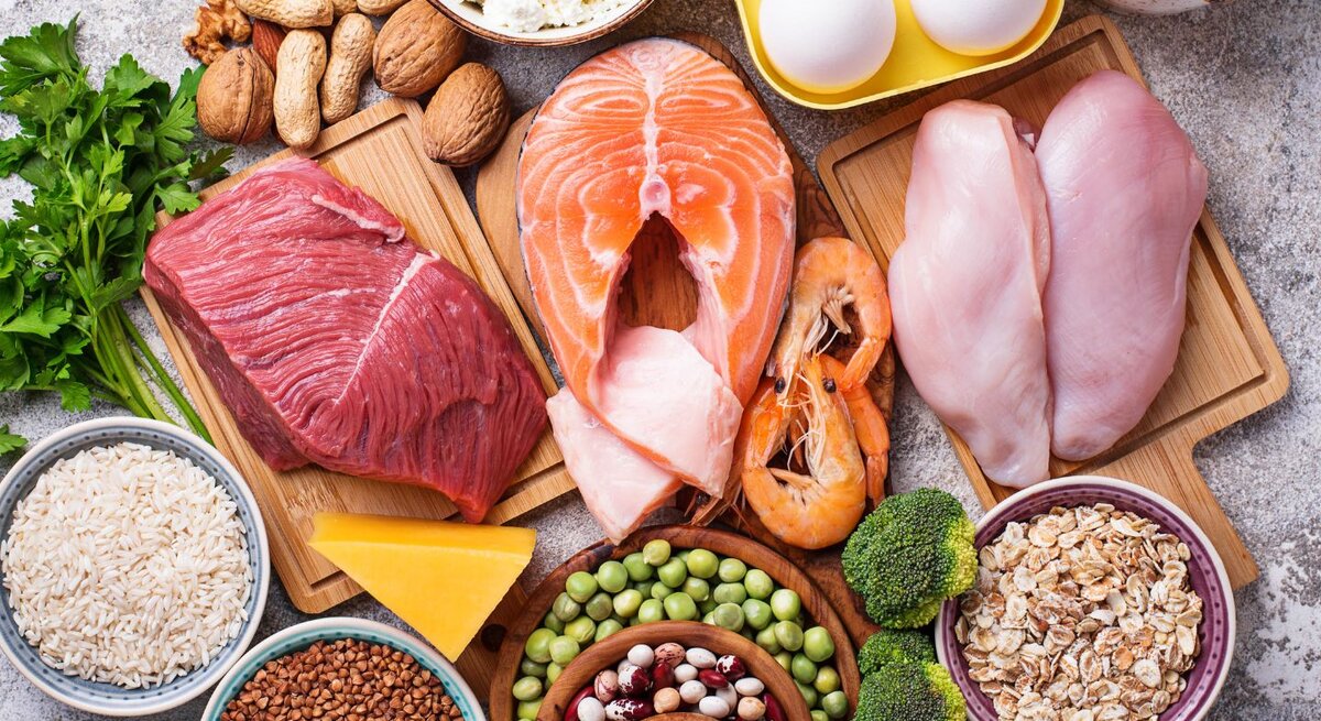Auswahl proteinreicher Lebensmittel: rohes Rindfleisch, Lachs, Hähnchenbrust, Eier, Garnelen, Hüttenkäse, Nüsse, Hülsenfrüchte, Erbsen, Reis, Haferflocken, Buchweizen, Brokkoli, Käse und Milch, arrangiert auf Brettern und in Schalen.