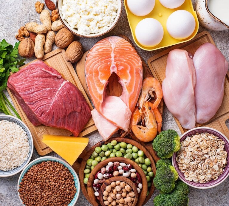 Auswahl proteinreicher Lebensmittel: rohes Rindfleisch, Lachs, Hähnchenbrust, Eier, Garnelen, Hüttenkäse, Nüsse, Hülsenfrüchte, Erbsen, Reis, Haferflocken, Buchweizen, Brokkoli, Käse und Milch, arrangiert auf Brettern und in Schalen. | © Getty Images/ yulka3ice via Canva.com