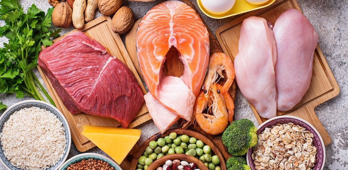 Auswahl proteinreicher Lebensmittel: rohes Rindfleisch, Lachs, Hähnchenbrust, Eier, Garnelen, Hüttenkäse, Nüsse, Hülsenfrüchte, Erbsen, Reis, Haferflocken, Buchweizen, Brokkoli, Käse und Milch, arrangiert auf Brettern und in Schalen. | © Getty Images/ yulka3ice via Canva.com