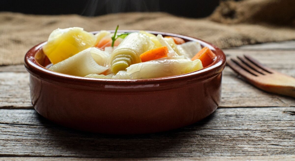 Nahaufnahme einer dampfenden Gemüsesuppe in einer runden, braunen Keramikschale auf einem rustikalen Holztisch: sichtbar sind große Stücke Lauch, Zwiebel und Möhren in klarer Brühe, oben ein kleines grünes Kräuterblatt. Der Hintergrund ist unscharf mit grobem Leinentuch; rechts liegt eine hölzerne Gabel. Warmes, natürliches Licht und gemütliche, erdige Farbtöne.