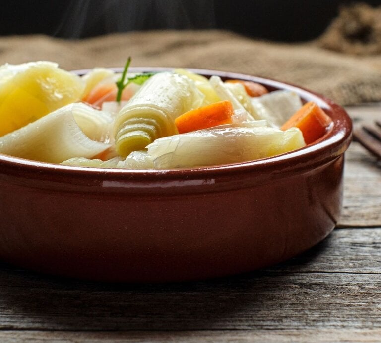 Nahaufnahme einer dampfenden Gemüsesuppe in einer runden, braunen Keramikschale auf einem rustikalen Holztisch: sichtbar sind große Stücke Lauch, Zwiebel und Möhren in klarer Brühe, oben ein kleines grünes Kräuterblatt. Der Hintergrund ist unscharf mit grobem Leinentuch; rechts liegt eine hölzerne Gabel. Warmes, natürliches Licht und gemütliche, erdige Farbtöne. | © Getty Images/ Alvaro Fernandez Echeverria via Canva.com