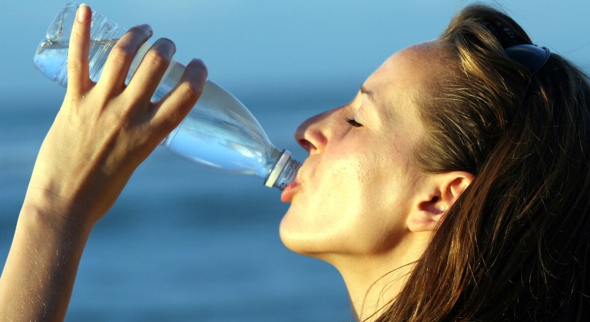 Eine Frau mit langen, dunkelbraunen Haaren trinkt bei Hitze aus einer transparenten Plastikflasche. Ihr Gesicht ist leicht nach oben geneigt, die Augen sind geschlossen. Die Haut wirkt erhitzt und leicht schweißbedeckt. Im Hintergrund ist unscharf ein blauer Himmel oder ein Gewässer zu sehen. Das Bild vermittelt Erfrischung und Flüssigkeitszufuhr bei sommerlichen Temperaturen.