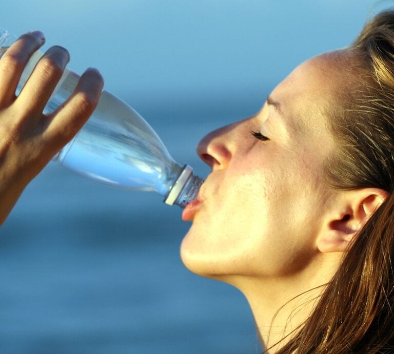 Eine Frau mit langen, dunkelbraunen Haaren trinkt bei Hitze aus einer transparenten Plastikflasche. Ihr Gesicht ist leicht nach oben geneigt, die Augen sind geschlossen. Die Haut wirkt erhitzt und leicht schweißbedeckt. Im Hintergrund ist unscharf ein blauer Himmel oder ein Gewässer zu sehen. Das Bild vermittelt Erfrischung und Flüssigkeitszufuhr bei sommerlichen Temperaturen. | © Adobe Stock/ Photo_Ma