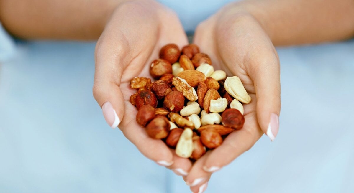 Nahaufnahme von zwei geöffneten Händen, die eine gemischte Portion Nüsse halten. Zu sehen sind unter anderem Mandeln, Haselnüsse, Cashews und Walnüsse. Der Hintergrund ist unscharf und hellblau, wodurch die Nüsse im Mittelpunkt stehen.“