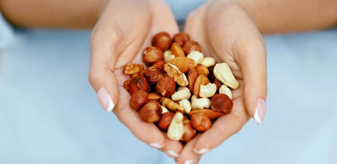 Nahaufnahme von zwei geöffneten Händen, die eine gemischte Portion Nüsse halten. Zu sehen sind unter anderem Mandeln, Haselnüsse, Cashews und Walnüsse. Der Hintergrund ist unscharf und hellblau, wodurch die Nüsse im Mittelpunkt stehen.“ | © Getty Images/ puhhha via Canva.com