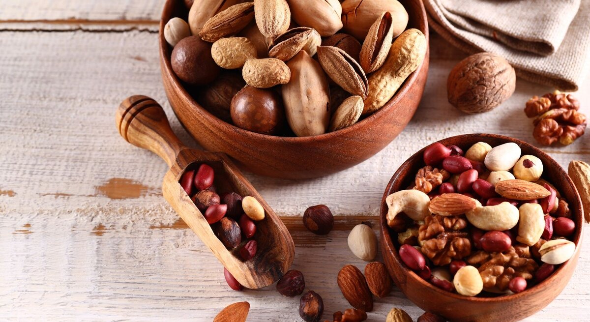 Zwei Holzschalen mit gemischten Nüssen und Erdnüssen auf einem hellen Holztisch. Zu sehen sind unter anderem Mandeln, Walnüsse, Cashews, Pistazien und Haselnüsse, daneben liegt eine kleine Holzschaufel mit weiteren Nüssen.