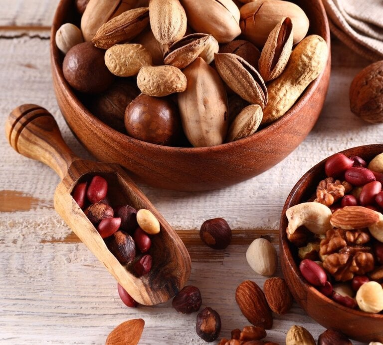 Zwei Holzschalen mit gemischten Nüssen und Erdnüssen auf einem hellen Holztisch. Zu sehen sind unter anderem Mandeln, Walnüsse, Cashews, Pistazien und Haselnüsse, daneben liegt eine kleine Holzschaufel mit weiteren Nüssen. | © Olga Kriger via Canva.com