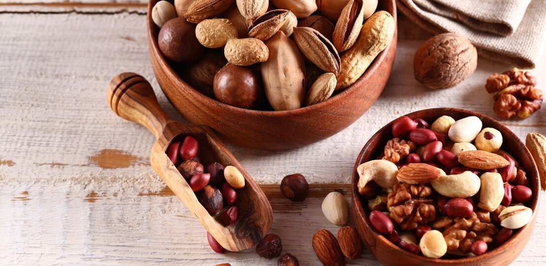 Zwei Holzschalen mit gemischten Nüssen und Erdnüssen auf einem hellen Holztisch. Zu sehen sind unter anderem Mandeln, Walnüsse, Cashews, Pistazien und Haselnüsse, daneben liegt eine kleine Holzschaufel mit weiteren Nüssen. | © Olga Kriger via Canva.com