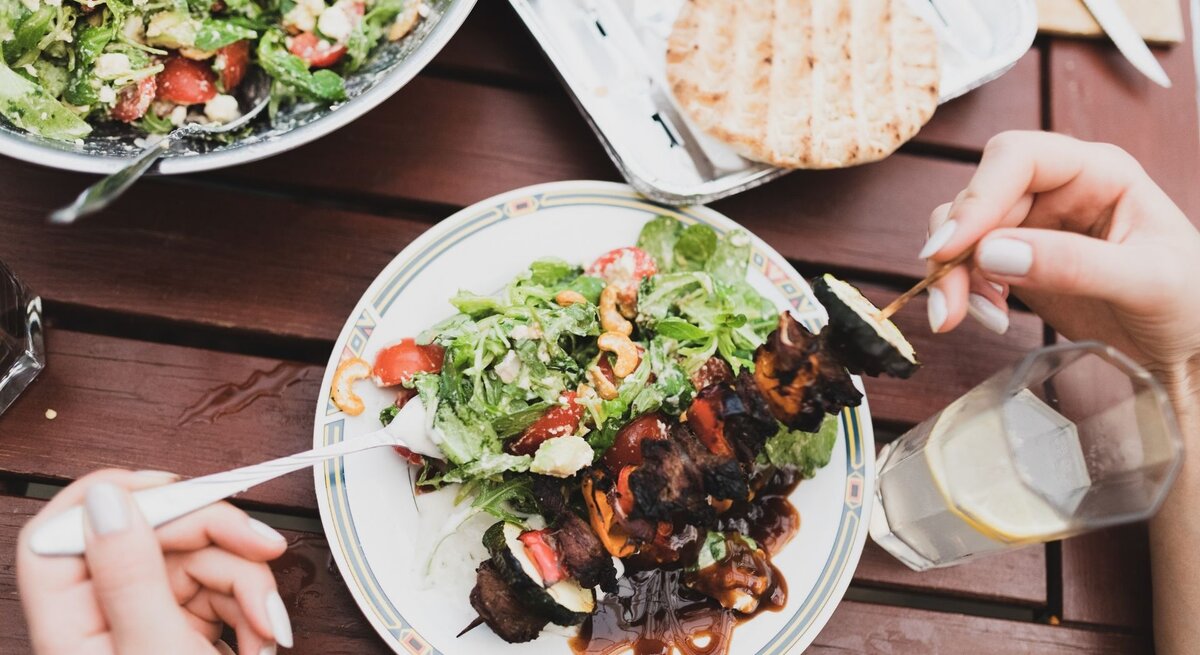 Vogelperspektive auf einen sommerlich gedeckten Holztisch. Auf einem gemusterten Teller in der Mitte ist ein grüner Salat mit Tomaten, Cashewkernen und Schafskäse sowie ein gegrillter Fleisch-Gemüse-Spieß mit dunkler Sauce angerichtet. Zwei Hände sind beim Essen zu sehen: Eine hält eine Gabel, die andere einen Holzspieß. Im Hintergrund befinden sich eine große Salatschüssel, Fladenbrot und ein Glas Zitronenwasser.