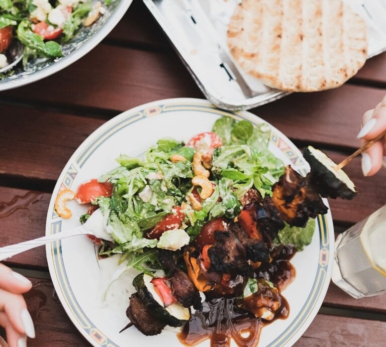 Vogelperspektive auf einen sommerlich gedeckten Holztisch. Auf einem gemusterten Teller in der Mitte ist ein grüner Salat mit Tomaten, Cashewkernen und Schafskäse sowie ein gegrillter Fleisch-Gemüse-Spieß mit dunkler Sauce angerichtet. Zwei Hände sind beim Essen zu sehen: Eine hält eine Gabel, die andere einen Holzspieß. Im Hintergrund befinden sich eine große Salatschüssel, Fladenbrot und ein Glas Zitronenwasser. | © Adobe Stock/ Extended License