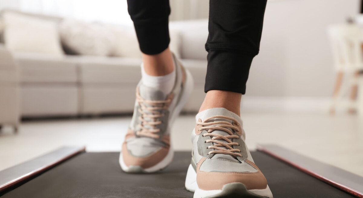 Füße in modernen Sneakers mit beige-grauen Details gehen auf einem schwarzen Walking Pad von Lidl in einem hellen Wohnzimmer