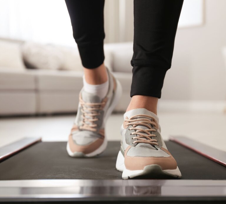Füße in modernen Sneakers mit beige-grauen Details gehen auf einem schwarzen Walking Pad von Lidl in einem hellen Wohnzimmer | © AdobeStock/ New Africa