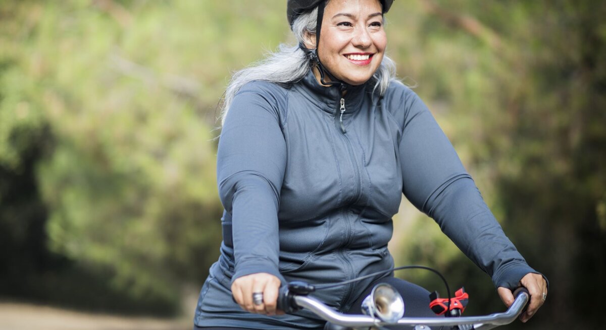 Eine fröhliche ältere Frau mit grau melierten Haaren fährt Rad in einem Freizeitoutfit. Sie trägt einen Fahrradhelm und ein langärmeliges Oberteil und lächelt, während sie auf einem Weg umgeben von grüner Vegetation radelt