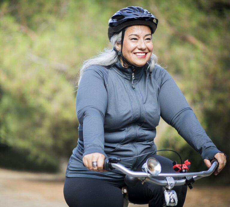Eine fröhliche ältere Frau mit grau melierten Haaren fährt Rad in einem Freizeitoutfit. Sie trägt einen Fahrradhelm und ein langärmeliges Oberteil und lächelt, während sie auf einem Weg umgeben von grüner Vegetation radelt | © gettyimages/	adamkaz