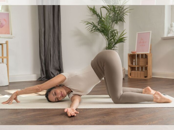 Eine Frau führt eine Yoga-Dehnübung aus, bei der sie mit einem Arm gestreckt auf einer Matte liegt, während sie den anderen Arm nach vorne ausstreckt. Im Hintergrund sind ein Zimmerpflanze, eine braune Holzablage und Wanddekorationen sichtbar. | © YogaEasy GmbH