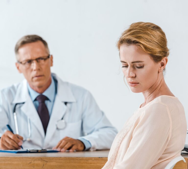 Eine nachdenklich blickende Frau sitzt mit gesenktem Kopf vor einem älteren Arzt in weißem Kittel an einem Schreibtisch. | © Adobe Stock / LIGHTFIELD STUDIOS