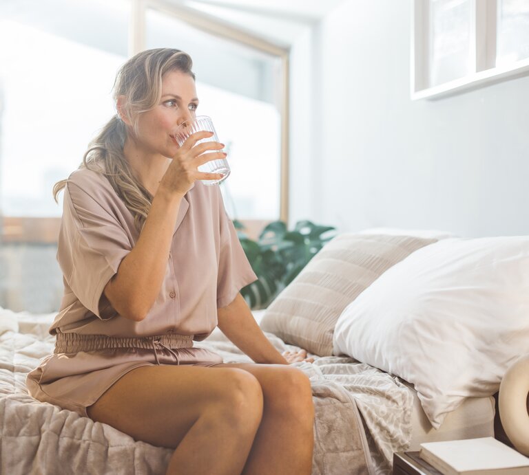 Eine Frau sitzt am Bettrand und trinkt aus einem Glas Wasser. | © Getty Images/svetikd