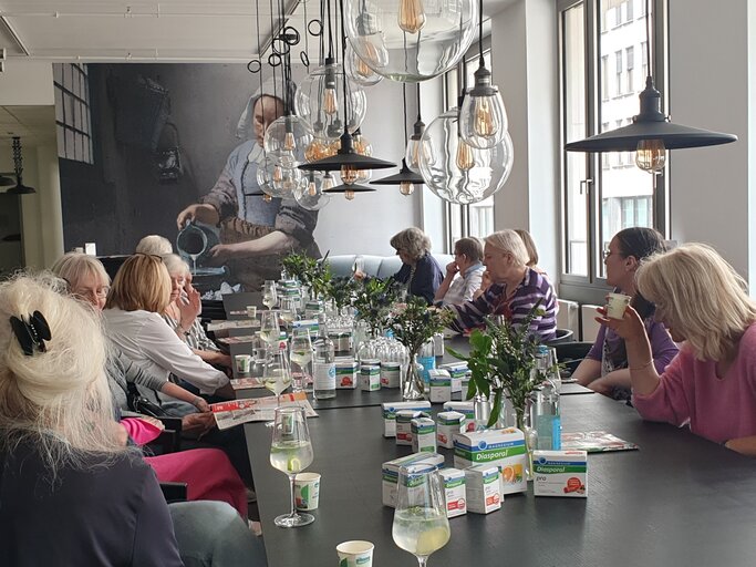 Eine Gruppe älterer Frauen sitzt tagsüber an langem Tisch mit Diasporal-Produkten, Zitronenwasser und Blumen unter Glaskugellampen. | © BILD der FRAU