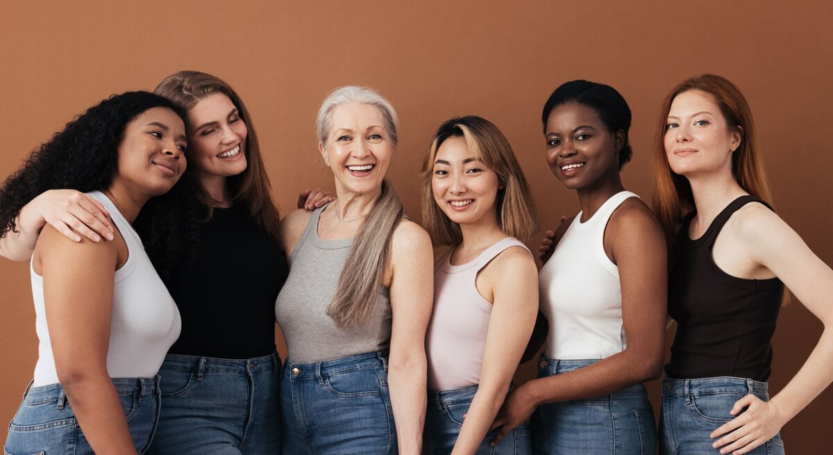 Sechs Frauen verschiedener Altersgruppen und Ethnien stehen eng umschlungen, lächelnd in Jeans und Tops vor braunem Hintergrund.