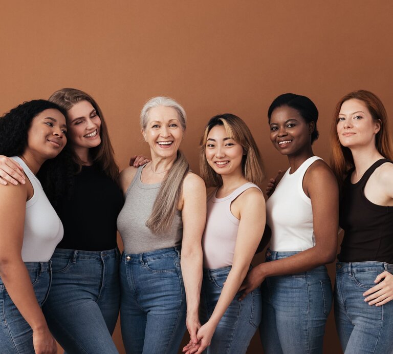 Sechs Frauen verschiedener Altersgruppen und Ethnien stehen eng umschlungen, lächelnd in Jeans und Tops vor braunem Hintergrund. | © Getty Images / Youngoldman