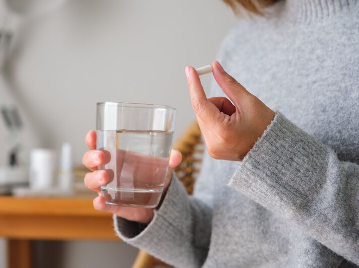 Eine Frau in grauem Wollpullover hält ein Wasserglas in der einen Hand und eine Magnesium-Tablette. Sie hat ihre Periode. | © Adobe Stock/ Farknot Architect