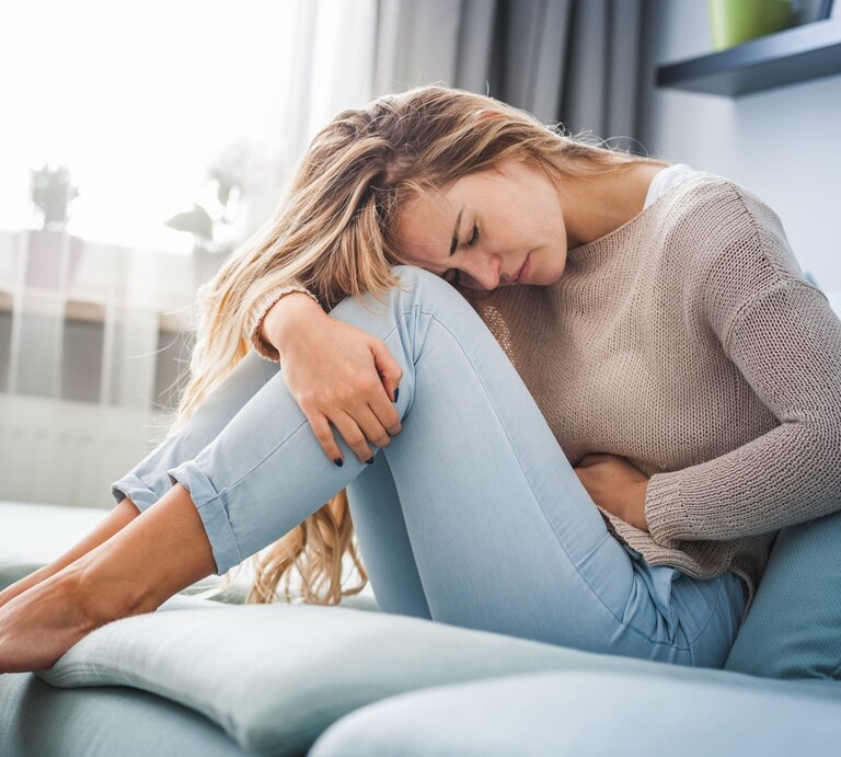 Eine Frau mit langen blonden Haaren sitzt mit schmerzverzerrtem Gesicht auf einem blauen Sofa und hält sich den Bauch. | © Adobe Stock/ leszekglasner