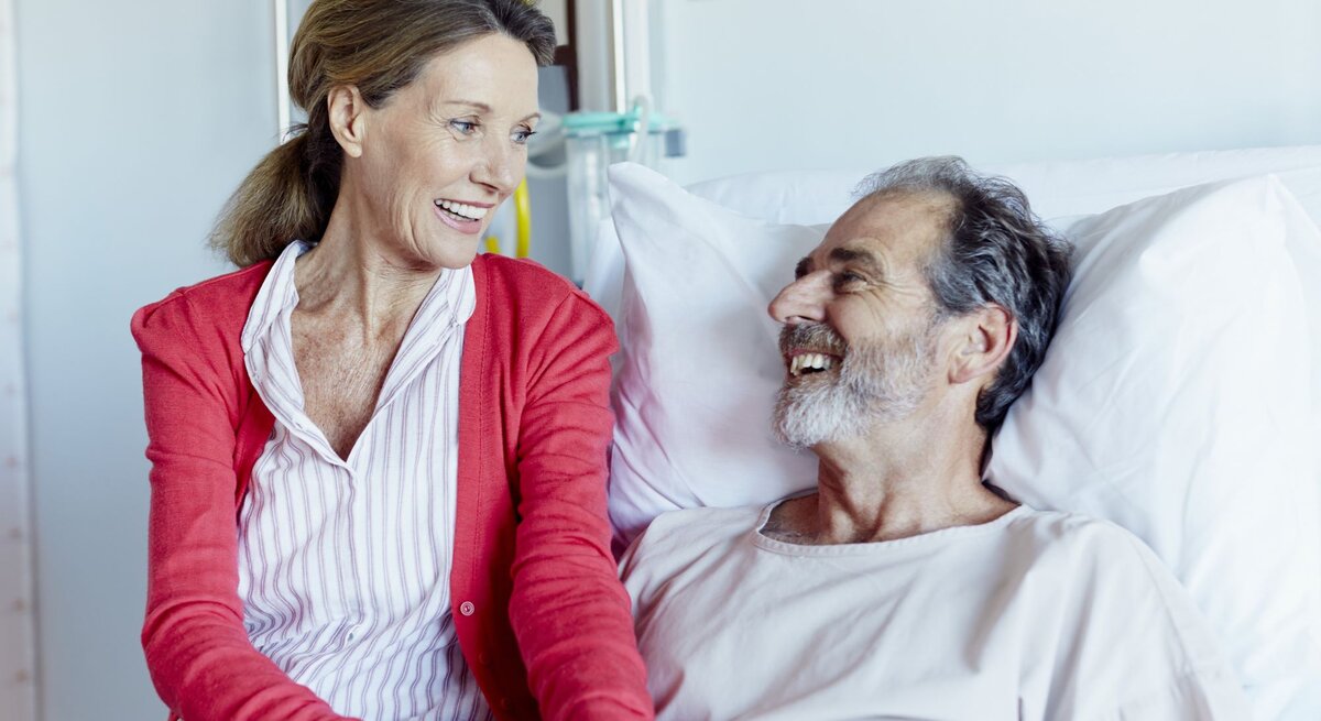 Frau, die an der Seite eines Mannes sitzt, der in einem Krankenhausbett liegt. Beide lächeln sich an und wirken entspannt und glücklich. 