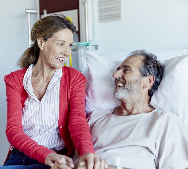 Frau, die an der Seite eines Mannes sitzt, der in einem Krankenhausbett liegt. Beide lächeln sich an und wirken entspannt und glücklich.  | © iStock