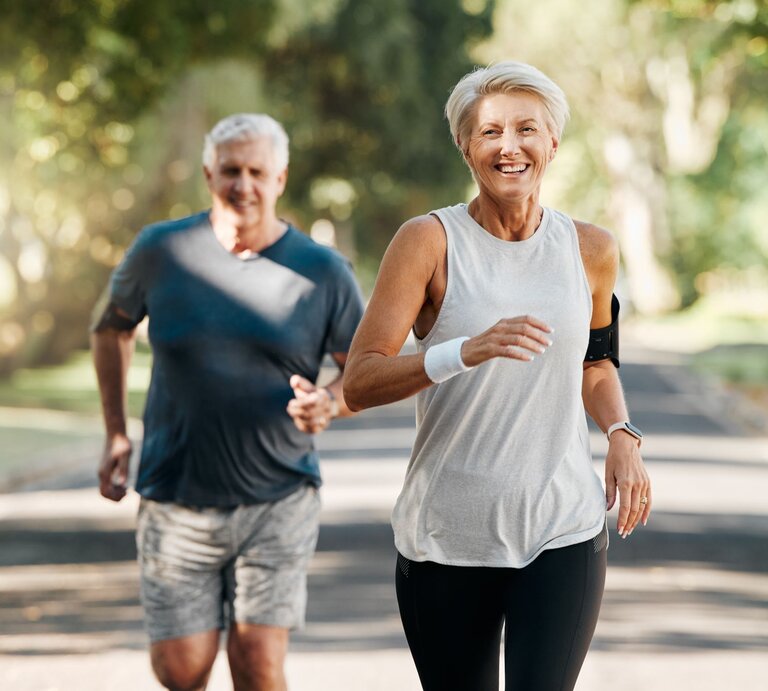 Älteres Paar, das gemeinsam im Freien joggt. Beide sehen glücklich und entspannt aus, während sie durch eine von Bäumen gesäumte Straße laufen. | © Adobe Stock