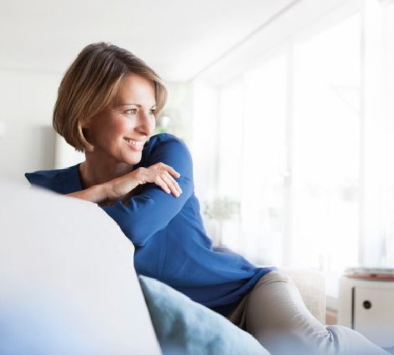 Frau mittleren Alters sitzt auf Sofa und schaut zur Seite | © Getty Images/Westend61