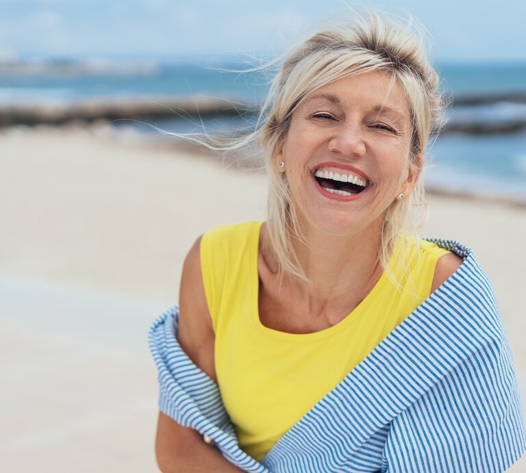Dame mittleren Alters steht lachend am Strand | © stock.adobe.com – contrastwerkstatt