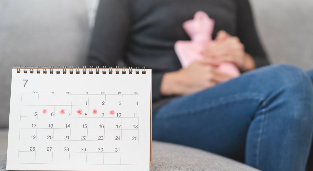 Das Bild zeigt eine Person, die auf einem Sofa sitzt und eine Wärmflasche an den Unterleib drückt, ein Zeichen für Menstruationsbeschwerden. Im Vordergrund steht ein Kalender auf einem Tisch, mit einigen Tagen markiert, was darauf hindeuten könnte, dass die Person ihre Menstruationszyklen verfolgt. Der Kalender fokussiert auf den Monat Juli, und die markierten Tage scheinen die erwartete Periode darzustellen.