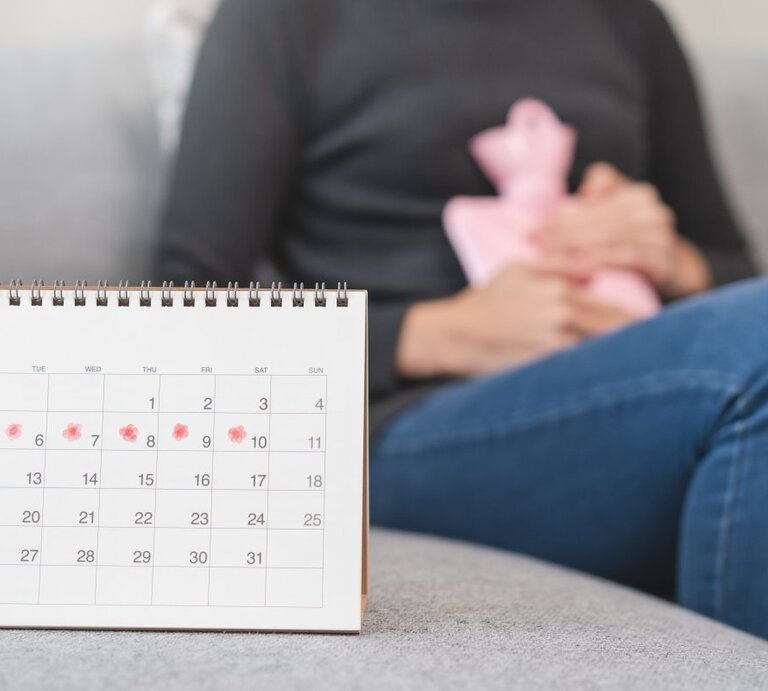 Das Bild zeigt eine Person, die auf einem Sofa sitzt und eine Wärmflasche an den Unterleib drückt, ein Zeichen für Menstruationsbeschwerden. Im Vordergrund steht ein Kalender auf einem Tisch, mit einigen Tagen markiert, was darauf hindeuten könnte, dass die Person ihre Menstruationszyklen verfolgt. Der Kalender fokussiert auf den Monat Juli, und die markierten Tage scheinen die erwartete Periode darzustellen. | © shutterstock/Pormezz