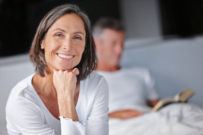 Reife Frau sitzt auf Bett und schaut in die Kamera während ihr Mann im Hintergrund liest | ©  iStock/laflor