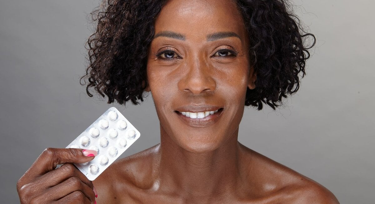 Das Bild zeigt eine lächelnde Frau mit kurzen lockigen Haaren, die eine Blisterpackung mit weißen Tabletten in der Hand hält. Sie präsentiert die Packung direkt in die Kamera, während ihr Gesicht und ihre Schultern scharf im Vordergrund zu sehen sind. Der Hintergrund ist neutral und ermöglicht eine klare Fokussierung auf die Person und die Tablettenpackung. Die Frau scheint zuversichtlich und könnte die Bedeutung von Verhütung oder Gesundheitsvorsorge während der Wechseljahre symbolisieren.
