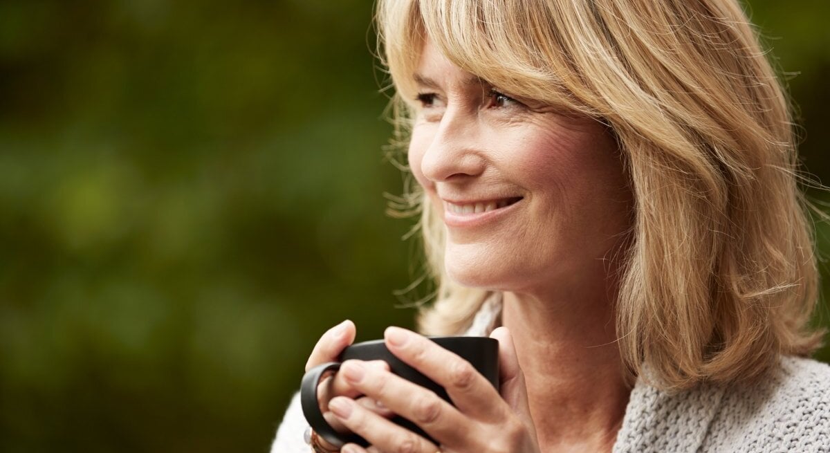 Das Foto zeigt eine reife Frau mit blonden Haaren, die lächelnd in die Ferne schaut, während sie eine dunkle Teetasse in ihren Händen hält. Sie trägt einen gemütlichen grauen Strickpullover und scheint sich in einem entspannten Moment im Freien zu befinden, mit einem unscharfen grünen Hintergrund, der auf einen Garten oder Park hindeuten könnte. Die sanfte Mimik der Frau strahlt Ruhe und Zufriedenheit aus.