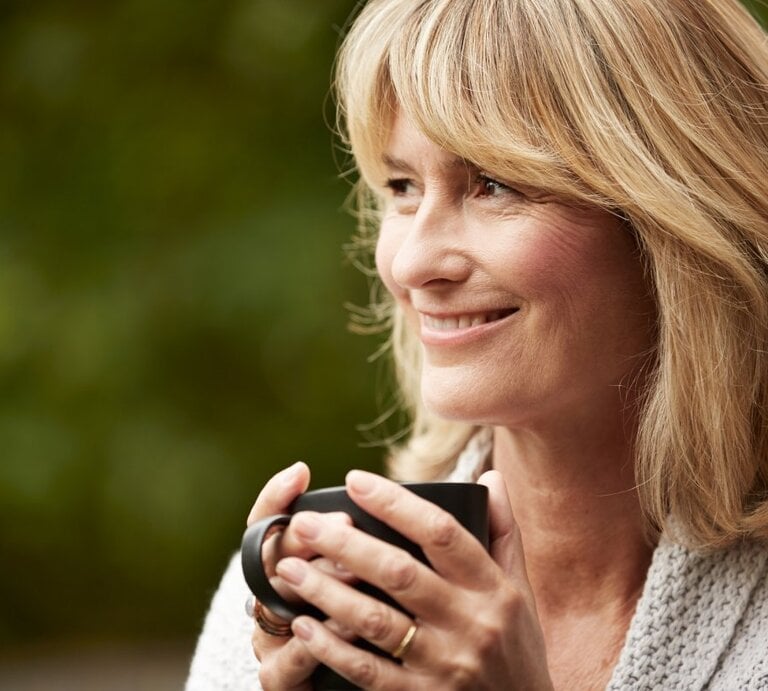 Das Foto zeigt eine reife Frau mit blonden Haaren, die lächelnd in die Ferne schaut, während sie eine dunkle Teetasse in ihren Händen hält. Sie trägt einen gemütlichen grauen Strickpullover und scheint sich in einem entspannten Moment im Freien zu befinden, mit einem unscharfen grünen Hintergrund, der auf einen Garten oder Park hindeuten könnte. Die sanfte Mimik der Frau strahlt Ruhe und Zufriedenheit aus. | © iStock/kupicoo