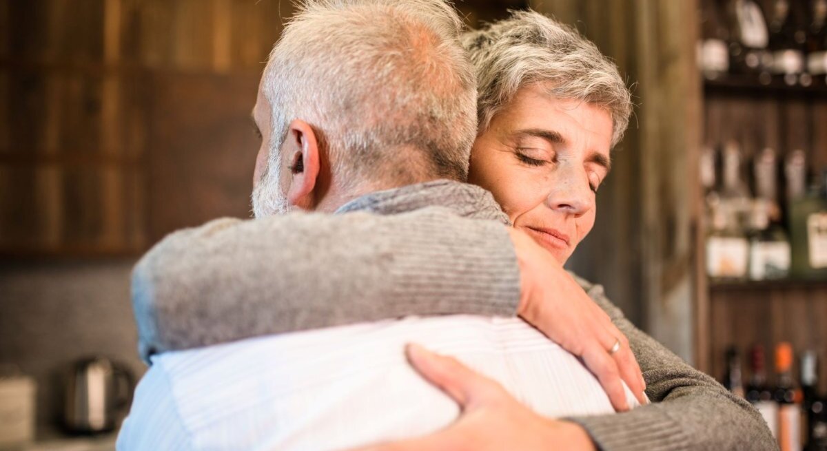 Das Foto zeigt zwei ältere Personen, die sich in einer herzlichen Umarmung befinden. Die Frau, die in die Kamera blickt, hat kurzes graues Haar und ein geschlossenes, friedliches Lächeln, während der Mann ihr seine Zuneigung zeigt, indem er sie fest hält. Das Paar scheint sich in einer häuslichen oder gemütlichen Umgebung zu befinden, was durch unscharfe Hintergrundelemente wie Regale mit verschiedenen Gegenständen angedeutet wird. Das Bild strahlt Wärme, Liebe und Fürsorge aus.