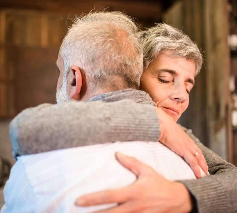 Das Foto zeigt zwei ältere Personen, die sich in einer herzlichen Umarmung befinden. Die Frau, die in die Kamera blickt, hat kurzes graues Haar und ein geschlossenes, friedliches Lächeln, während der Mann ihr seine Zuneigung zeigt, indem er sie fest hält. Das Paar scheint sich in einer häuslichen oder gemütlichen Umgebung zu befinden, was durch unscharfe Hintergrundelemente wie Regale mit verschiedenen Gegenständen angedeutet wird. Das Bild strahlt Wärme, Liebe und Fürsorge aus. | © Getty Images / franckreporter