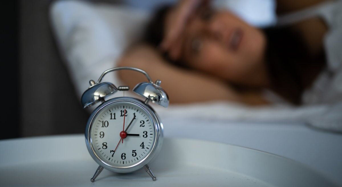 Das Foto zeigt einen klassischen Wecker im Vordergrund, der auf einem Nachttisch steht, mit einer unscharfen Person im Hintergrund, die im Bett liegt und sich anscheinend mit Schlafschwierigkeiten herumschlägt. Die Person hält sich eine Hand an die Stirn in einer Geste von Frustration oder Erschöpfung. Der Fokus auf den Wecker unterstreicht das Thema Schlaflosigkeit und die damit verbundenen Herausforderungen.
