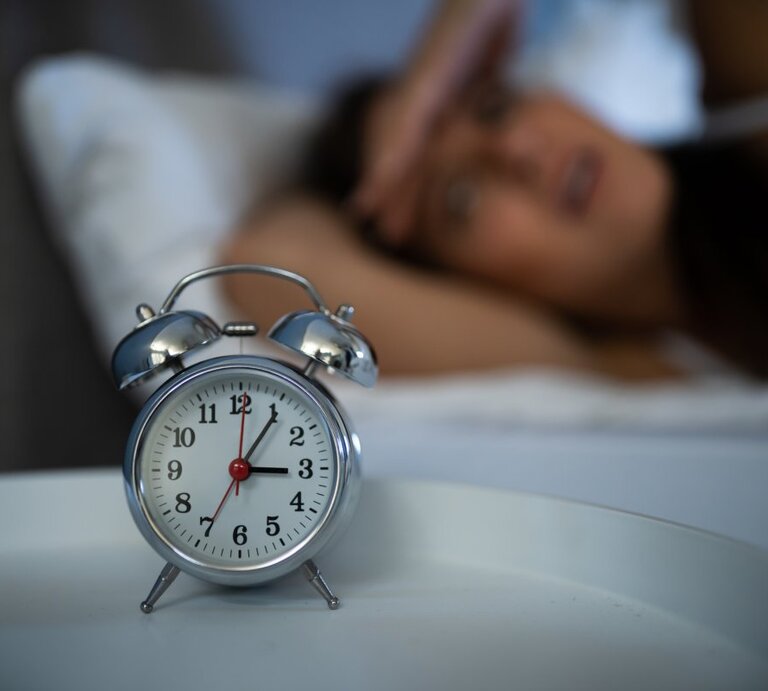 Das Foto zeigt einen klassischen Wecker im Vordergrund, der auf einem Nachttisch steht, mit einer unscharfen Person im Hintergrund, die im Bett liegt und sich anscheinend mit Schlafschwierigkeiten herumschlägt. Die Person hält sich eine Hand an die Stirn in einer Geste von Frustration oder Erschöpfung. Der Fokus auf den Wecker unterstreicht das Thema Schlaflosigkeit und die damit verbundenen Herausforderungen. | © Getty Images / AndreyPopov