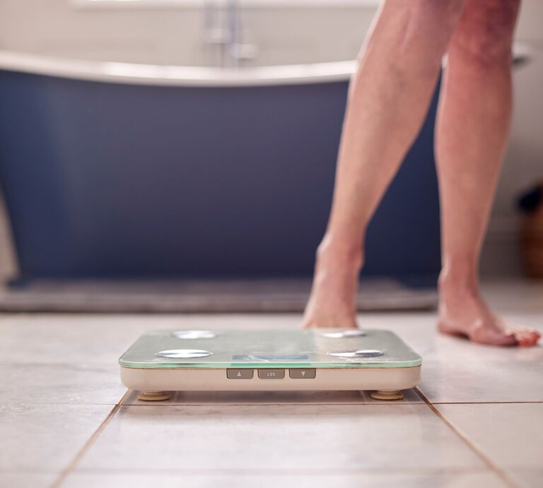 Das Foto zeigt die untere Hälfte einer Person, die im Begriff steht, auf eine Personenwaage im Badezimmer zu steigen. Man sieht nur die Beine und Füße der Person, die Waage ist im Vordergrund des Bildes. Im Hintergrund ist ein Teil einer blauen Badewanne und ein geflochtener Wäschekorb zu erkennen. Der Fokus liegt auf der Aktion des Wiegen als Teil der Gesundheits- und Körperpflegeroutine. | © Adobe Stock/ Monkey Business