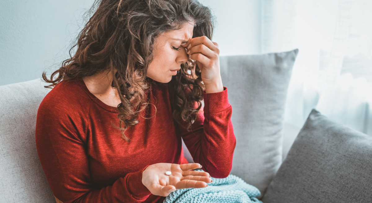 Junge Frau mit langen braunen Locken sitzt erschöpft auf dem Sofa, hält Tabletten in der Hand und reibt sich die Stirn.