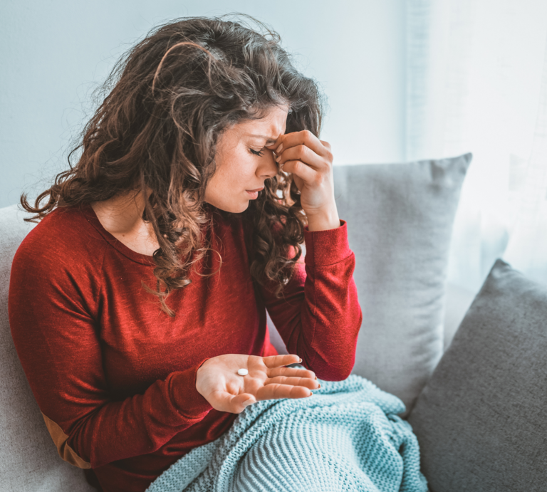 Junge Frau mit langen braunen Locken sitzt erschöpft auf dem Sofa, hält Tabletten in der Hand und reibt sich die Stirn. | © Canva