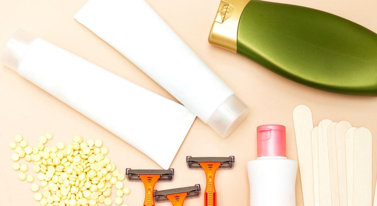 Eine Reihe von Hygieneprodukten auf einem Tisch, darunter gelbes Wachs in einem Behälter, ein Holzstäbchen, ein Rasierer, sowie Flaschen mit Intimlotion und -shampoo, alle arrangiert für die persönliche Pflege.
