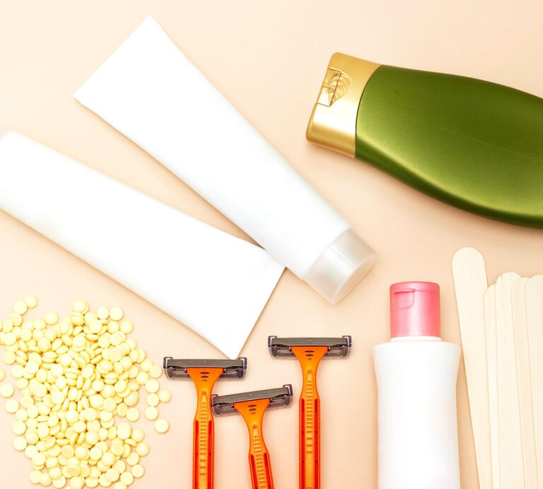 Eine Reihe von Hygieneprodukten auf einem Tisch, darunter gelbes Wachs in einem Behälter, ein Holzstäbchen, ein Rasierer, sowie Flaschen mit Intimlotion und -shampoo, alle arrangiert für die persönliche Pflege. | © Adobe Stock/hadjanebia