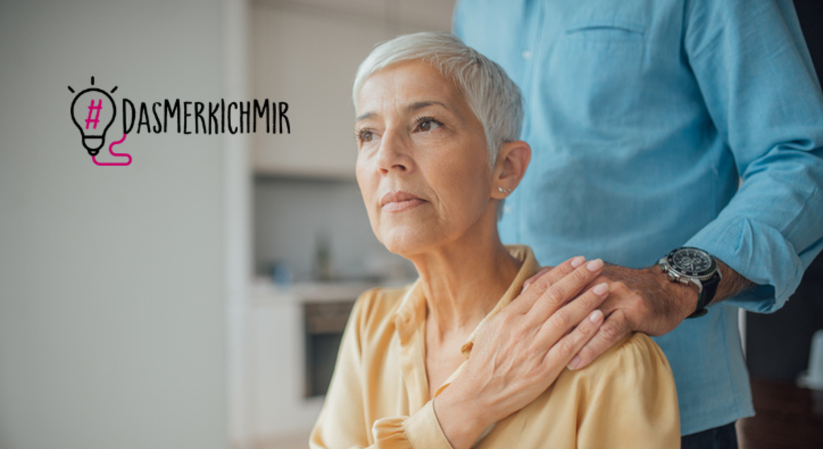 Eine ältere Frau mit kurzen grauen Haaren schaut nachdenklich zur Seite. Sie trägt eine dunkelgelbe Bluse. Hinter ihr steht jemand, der nur teilweise im Bild zu sehen ist und dessen Gesicht nicht erkennbar ist. Die Person legt der Frau tröstend eine Hand auf ihre Schulter. Die Person trägt ein blaues Hemd und eine Uhr. Neben ihnen ist der Hashtag "#DAS MERK ICH MIR" mit einer Glühbirnen-Grafik zu sehen.