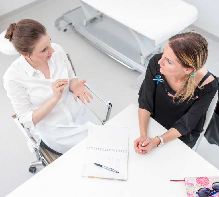 Ärztin im Gespräch mit einer Patientin, beide sitzen an einem Tisch, Perspektive von oben. | © Canva