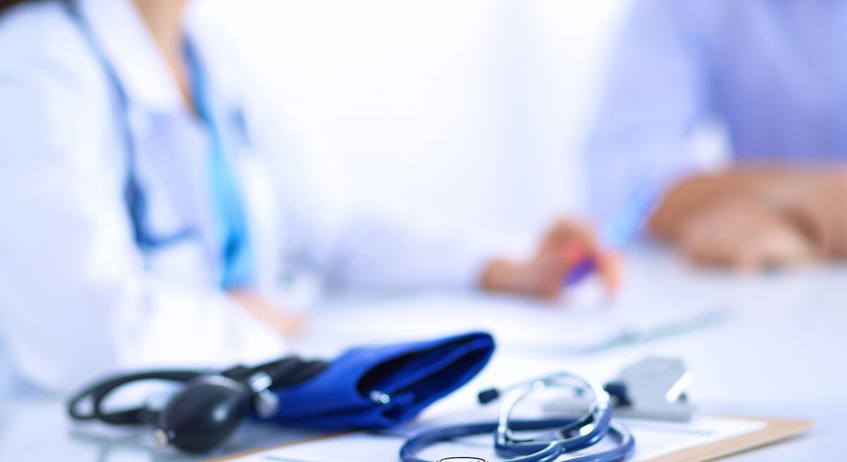 Beratungsgespräch in Arztpraxis, weibliche Ärztin mit blauem Stethoskop spricht mit Patient.