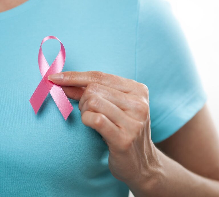Eine Frau in einem blauen T-Shirt hält eine rosa Schleife vor die linke Brust. | © gettyimages/	MarsBars